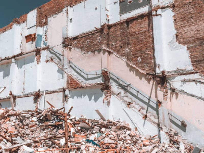 View of a brick wall of partly destroyed house during demolition works: railing on the wall, plaster, partially torn plaster, piles of construction waste in front, sunny day