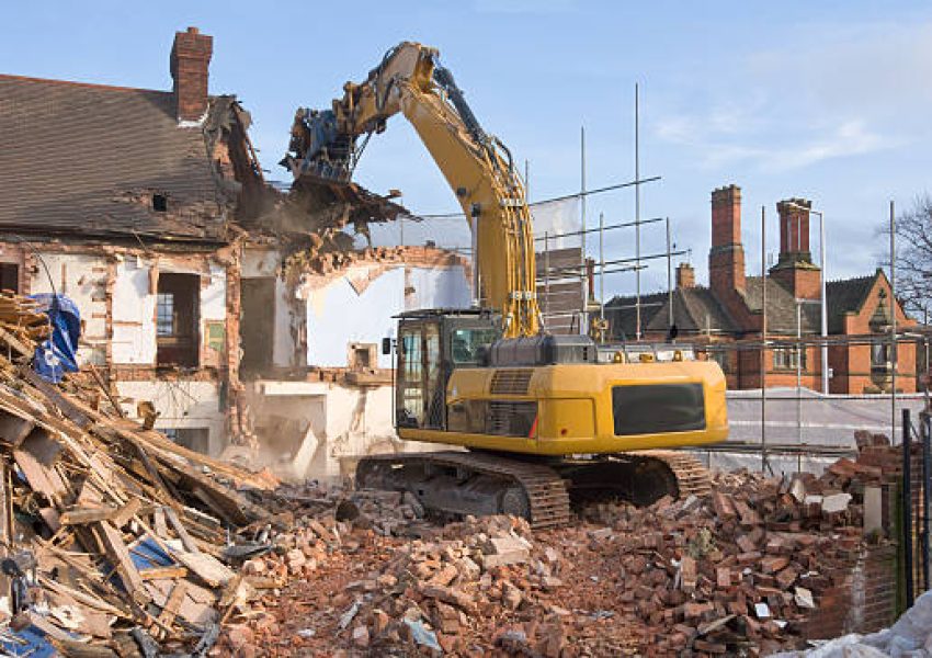 "A large tracked excavator machine fitted with a demolition tool, ripping apart old houses and buildings in a town centre for re-development."